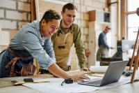 Des menuisiers souriants en tenue de travail utilisent un ordinateur portable et discutent dans leur atelier.